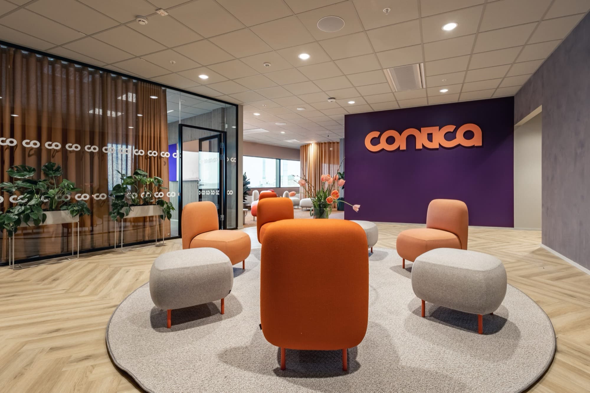 Entré med Conticas logotyp i orange på en lila vägg, runda sittpuffar i orange och grått på en rund matta och fiskbensparkett.