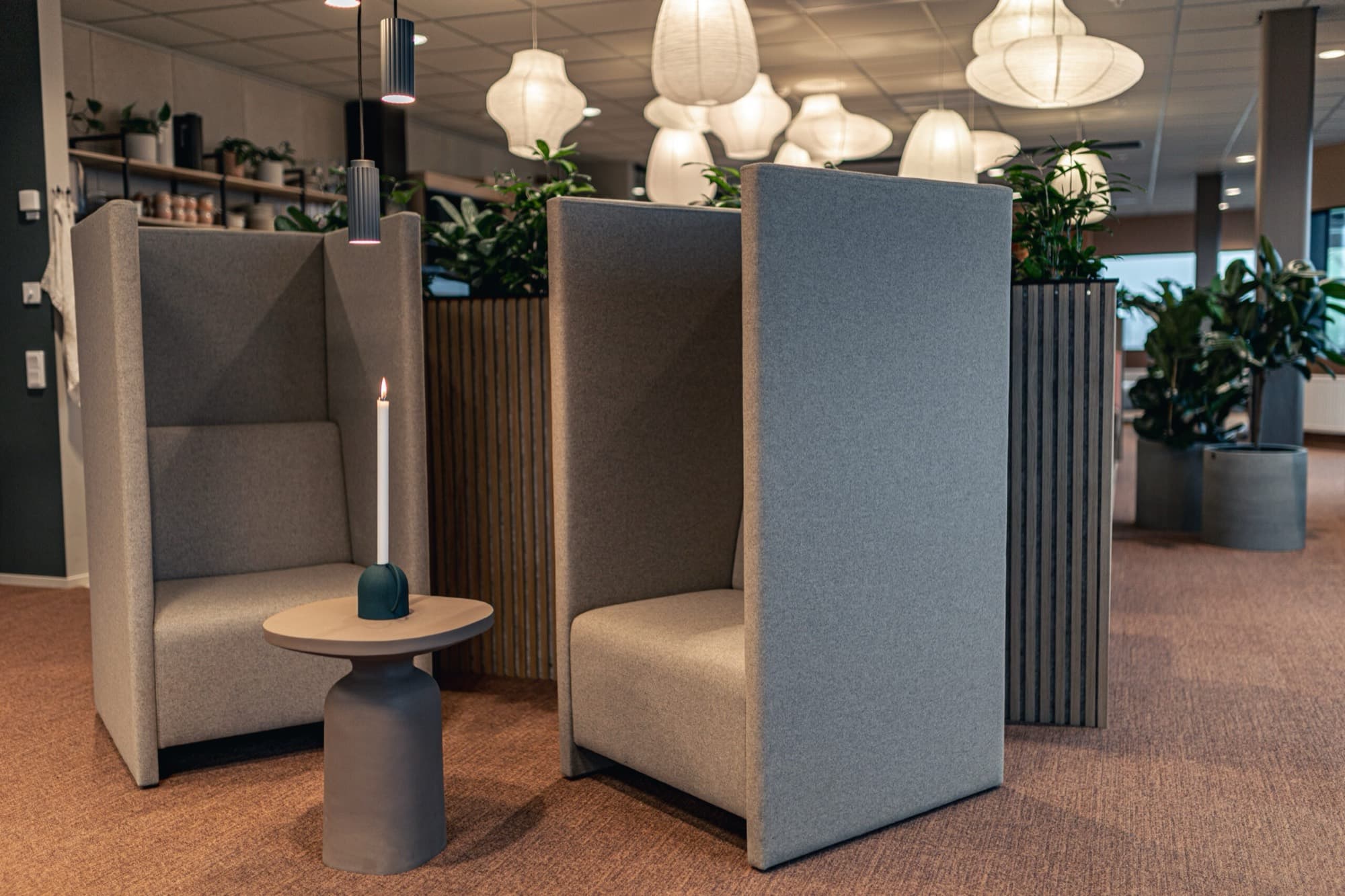 Två höga akustikfåtöljer i grått med ett litet bord emellan, i öppet landskap med rispapperslampor och växtavdelare.